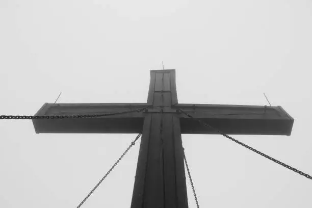 Wooden cross held by chains on top of Hochlantsch mountain in Fischbach Alps, Austria. A huge sign of the Christian faith lost in the white mist.