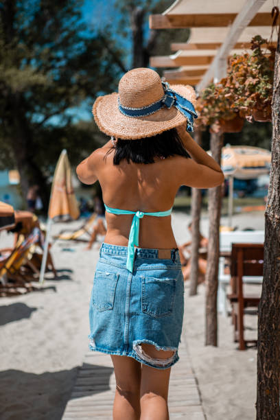 une vue arrière d’une jeune femme bronzée marchant sur la plage. elle porte une jupe en denim et un maillot de bain bleu, et elle porte un chapeau tricoté. - deck chair photos et images de collection