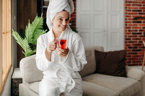 Beautiful girl in bath robe is holding a cup and smiling while resting at home