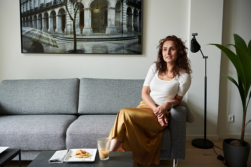 Pakistani woman with long brown hair is sitting in contemporary Danish apartment on a gray couch