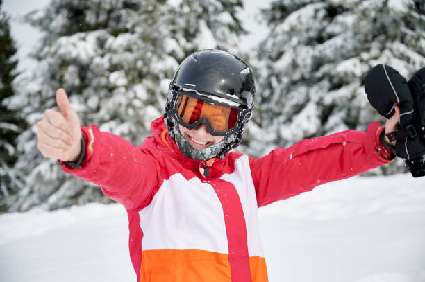 Cheerful female skier standing in winter mountain forest. Young woman in ski winter jacket and helmet looking at camera and smiling. Joyful female skier in ski goggles spreading hands to sides and giving thumbs up. Concept of joy and winter sport activities. thumbs up for skiing stock pictures, royalty-free photos & images