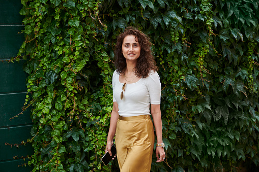 Portrait of woman with long brown hair dressed in golden skirt and white top. She is passing by some glreen plants hanging down from a wall.