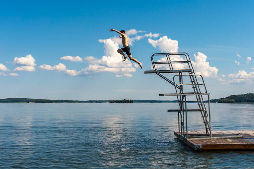 Side view of a teenage male jump diving from a diving tower with blue sky and horizon in the background. Hot summer afternoon at a bath place in lake Mälaren in Sweden, horizontal composition.