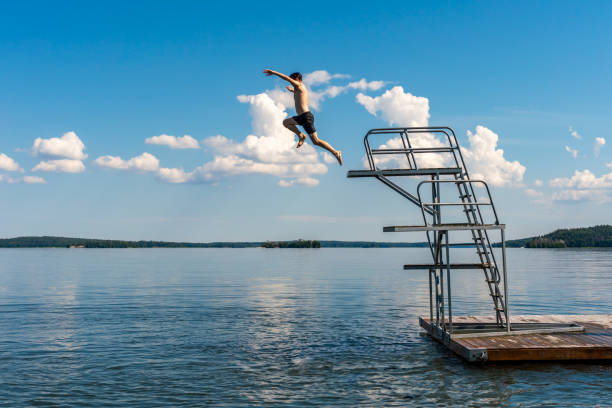 Side view of a teenage male jump diving from a diving tower with blue sky and horizon in the background. Side view of a teenage male jump diving from a diving tower with blue sky and horizon in the background. Hot summer afternoon at a bath place in lake Mälaren in Sweden, horizontal composition. swedish summer stock pictures, royalty-free photos & images