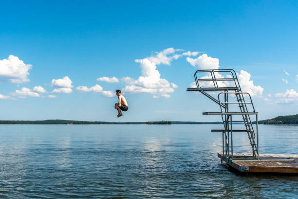 Side view of a teenage male jump diving from a diving tower with blue sky and horizon in the background. stock photo