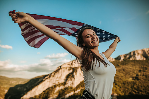 Beautiful young woman standing on top of a mountain and holding American flag on sunny summer day