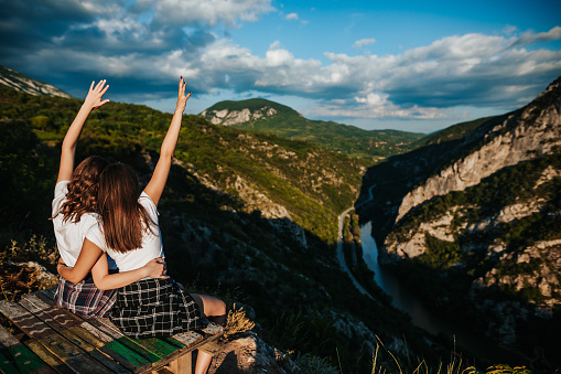 Two young female friends sitting on top of a mountain holding raised hands