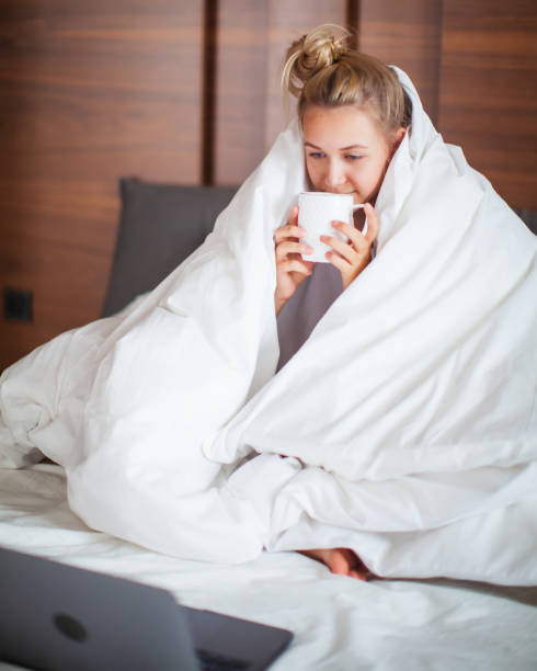 garota adolescente bonita debaixo de cobertor em cama branca no quarto acordou e bebeu chá com laptop. café da manhã na cama, manhã preguiçosa - five oclock tea - fotografias e filmes do acervo