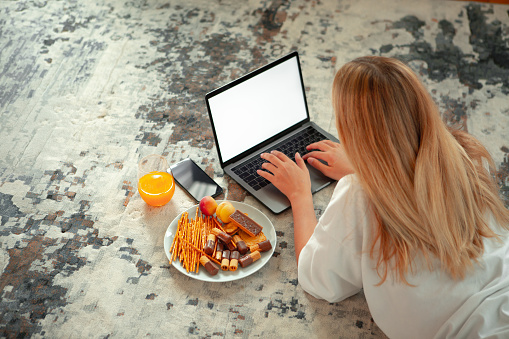Teen girl lying on floor with blank screen laptop watching TV series with delicious food and phone. High quality photo
