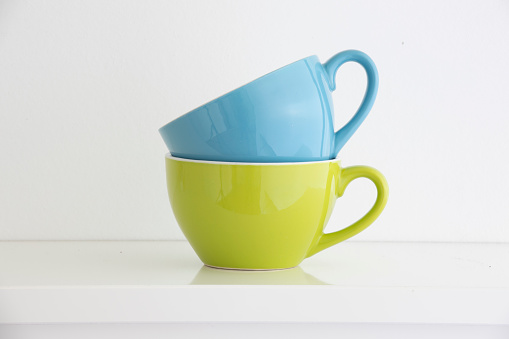 a blue and a green coffee cup stacked on a white shelf in front of a white wall, the handles are arranged on the right