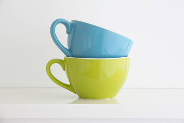 a blue and a green coffee cup stacked on a white shelf in front of a white wall, the handles are arranged on the left