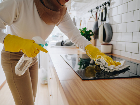 Young woman cleaning up the perfect surface of black ceramic kitchen stove and counter using gloves and mop.