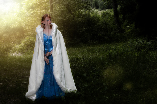 Young red-haired woman with elf ears, blue dress and white cape holds a sword in front of her face.