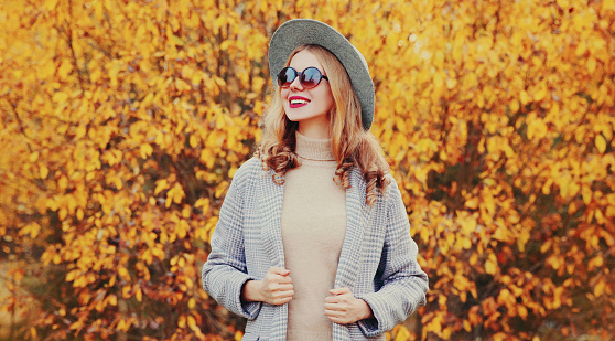 Autumn portrait of beautiful young woman wearing gray coat, round hat posing over yellow leaves background
