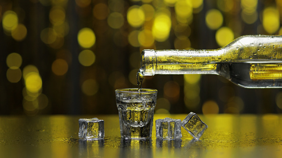 Bartender pouring up frozen vodka from bottle into shot glass with ice cubes against shiny gold party celebration background. Barman pour of cold transparent alcohol drink vodka tequila in shot-glass