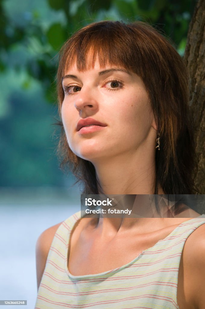 Serious look at camera Lateral head and shoulder portrait of a young brunette woman leaning against a tree, looking seriously at the camera. Germany Stock Photo