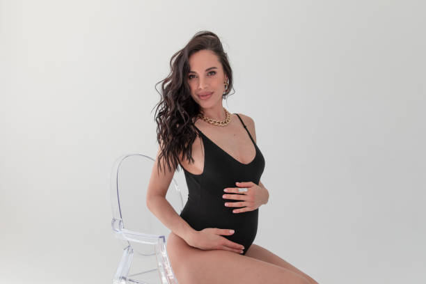 donna incinta di moda in bidysuit nero siede sulla sedia in studio bianco - women smiling body cheerful foto e immagini stock
