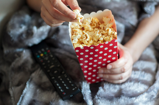 Woman hands eating popcorns wrapped on warm blanket and with tv remote control on lap