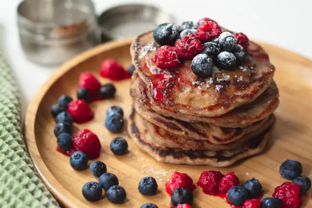 Photo of A heap of healthy vegan gluten free whole grain pancakes made with buckwheat flour topped with raspberries and blueberries with icing sugar sprinkled on top