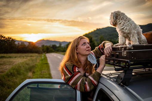 Corona virus - Covid 19 - A woman with a surgical mask travels with her dog
and flees into nature from an epidemic of the corona virus - Luggage on the roof of the car