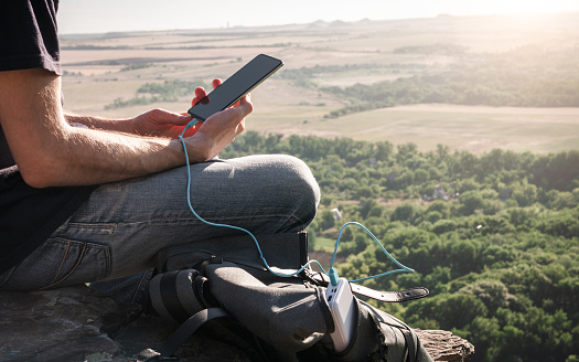 Man on a hike uses smartphone while charging from the power bank on the rock at dawn. Healthy lifestyle and communication
