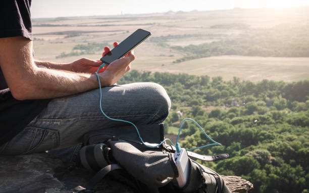 Man on a hike uses smartphone while charging from the power bank on the rock at dawn. Healthy lifestyle and communication. Man on a hike uses smartphone while charging from the power bank on the rock at dawn. Healthy lifestyle and communication portable power bank stock pictures, royalty-free photos & images