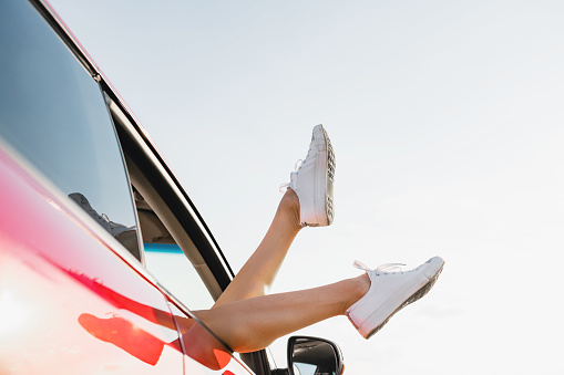 Playful girl sticking out feet in white sneakers from vehicle at sky background, happy traveling adventure.