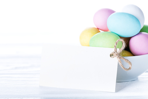 Colorful pastel easter eggs in bowl with blank paper tag for text on white wooden background, Happy Easter card