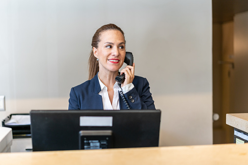 A middle aged female hotel manager is using telephone at front desk of the hotel