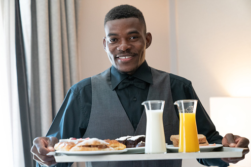 Room Service. Portrait of a happy young Afro-American waiter bringing breakfast to the hotel room.