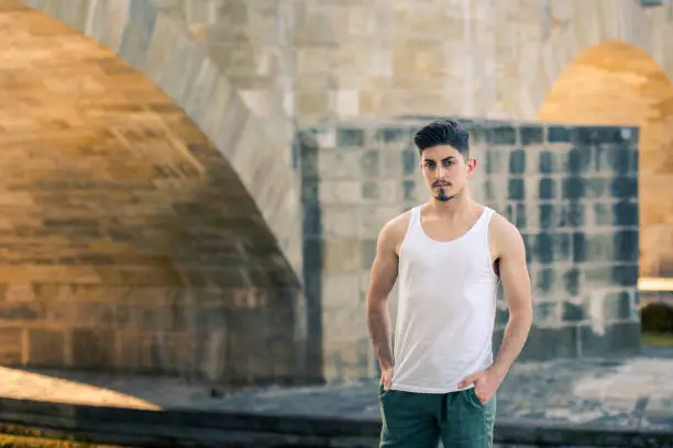 Urban portrait of young arabic man in summer outfit with standing next to arches of sunlit stone bridge on hot summer day looking at camera