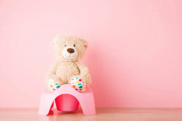 Photo of Brown teddy bear sitting on baby potty on floor. Front view. Closeup. Empty place for text on light pink background. Pastel color.