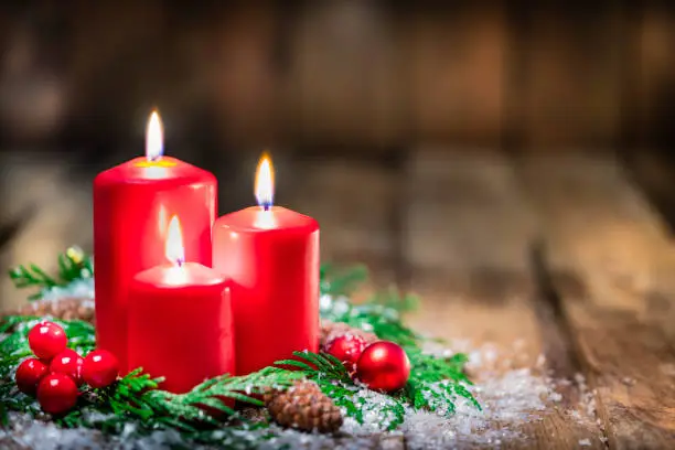 Christmas backgrounds: three red burning Christmas candles shot on rustic wooden table. The composition is at the left of an horizontal frame leaving useful copy space for text and/or logo at the right. Predominant colors are red and brown. High resolution 42Mp studio digital capture taken with Sony A7rII and Sony FE 90mm f2.8 macro G OSS lens