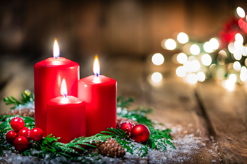 Christmas backgrounds: three red burning Christmas candles shot on rustic wooden table. The composition is at the left of an horizontal frame leaving useful copy space for text and/or logo at the right.\nString light and Christmas decoration are out of focus at background. Predominant colors are red and brown. High resolution 42Mp studio digital capture taken with Sony A7rII and Sony FE 90mm f2.8 macro G OSS lens