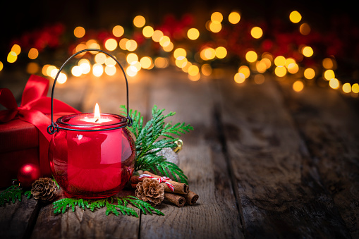 Christmas backgrounds: red burning Christmas candle shot on rustic wooden table. The composition is at the left of an horizontal frame leaving useful copy space for text and/or logo at the right.\nString light and Christmas decoration are out of focus at background. Predominant colors are red and brown. High resolution 42Mp studio digital capture taken with Sony A7rII and Sony FE 90mm f2.8 macro G OSS lens