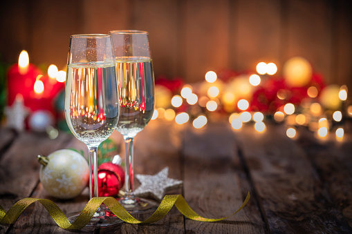 Two Champagne flutes on wooden Christmas table. Christmas candles and string lights are out of focus at background. The composition is at the left of an horizontal frame leaving useful copy space for text and/or logo at the right. Predominant colors are brown and yellow. High resolution 42Mp studio digital capture taken with Sony A7rII and Sony FE 90mm f2.8 macro G OSS lens