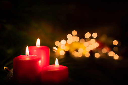 Christmas backgrounds: three red burning Christmas candles shot on rustic wooden table. The composition is at the left of an horizontal frame leaving useful copy space for text and/or logo at the right.\nString light and Christmas decoration are out of focus at background. Predominant colors are red and brown. High resolution 42Mp studio digital capture taken with SONY A7rII and Zeiss Batis 40mm F2.0 CF lens