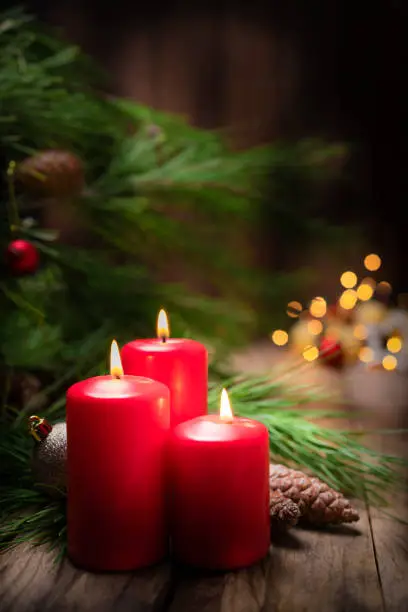 Christmas backgrounds: three red burning Christmas candles shot on rustic wooden table. The composition is at the left of an horizontal frame leaving useful copy space for text and/or logo at the right.
String light and Christmas decoration are out of focus at background. Predominant colors are red, green and brown. High resolution 42Mp studio digital capture taken with SONY A7rII and Zeiss Batis 40mm F2.0 CF lens