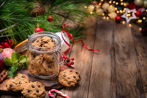 Homemade chocolate chips cookies jar shot on rustic wooden Christmas table. Christmas ornaments are around the cookies jar. A Christmas tree branch and string lights complete the composition. The composition is at the left of an horizontal frame leaving useful copy space for text and/or logo at the right. Predominant colors are brown and green. High resolution 42Mp studio digital capture taken with SONY A7rII and Zeiss Batis 40mm F2.0 CF lens