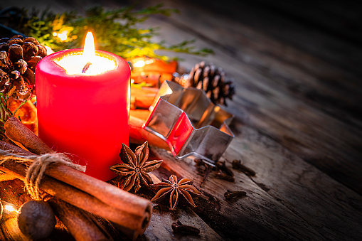 Christmas backgrounds: red burning Christmas candle shot on rustic wooden table. Cinnamon sticks, star anise, cloves and nutmeg are around the candle. The composition is at the left of an horizontal frame leaving useful copy space for text and/or logo at the right. Predominant colors are red and brown. High resolution 42Mp studio digital capture taken with Sony A7rII and Sony FE 90mm f2.8 macro G OSS lens