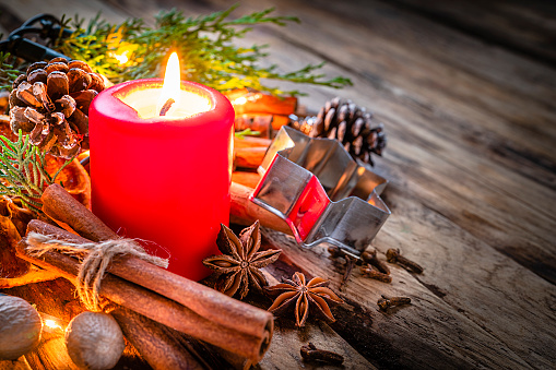 Christmas backgrounds: red burning Christmas candle shot on rustic wooden table. Cinnamon sticks, star anise, cloves and nutmeg are around the candle. The composition is at the left of an horizontal frame leaving useful copy space for text and/or logo at the right. Predominant colors are red and brown. High resolution 42Mp studio digital capture taken with Sony A7rII and Sony FE 90mm f2.8 macro G OSS lens