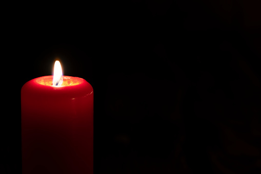 Red candle burning in the dark. Predominant colors are red, yellow and black. The composition is at the left of an horizontal frame leaving useful copy space for text and/or logo at the right. High resolution 42Mp studio digital capture taken with Sony A7rII and Sony FE 90mm f2.8 macro G OSS lens