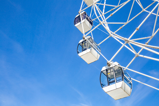 Big ferris wheel with white cabins in an amusement park against a blue sky during the sunny day