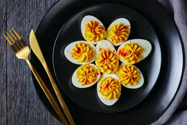 Classic deviled eggs filled with mustard, mayonnaise, white vinegar sprinkled with smoked paprika on black plate with golden cutlery on a dar wooden background, top view, flat lay