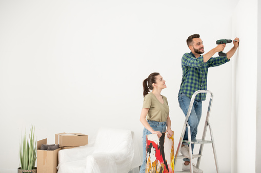 Positive young man standing on step ladder and using electric screwdriver while inserting screw for picture