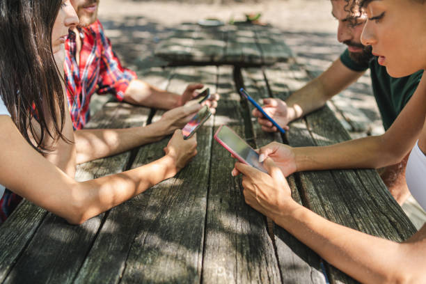 中庭の古い木製のテーブルの上にスマートフォンを使用している若者。夏の日曜日に携帯電話を使う友人たち。テクノロジーとソーシャルメディアのコンセプト。 - shadowed ストックフォトと画像