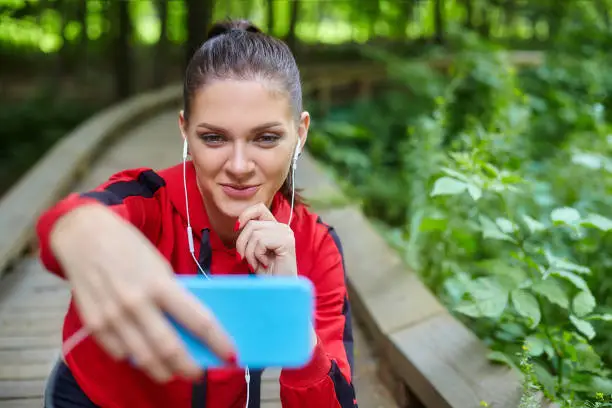 Distance learning concept. An attractive girl in sportswear sits on a wooden path in a forest park. Online lessons via smartphone.