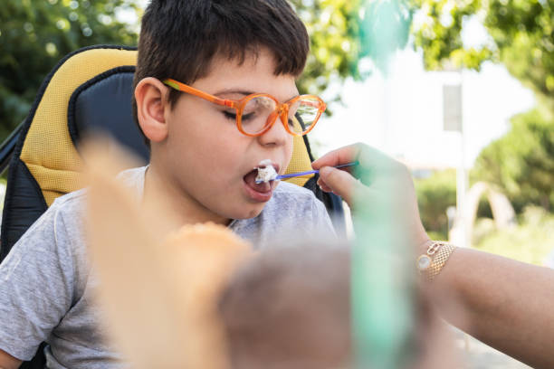 disabled child eating creamy ice cream with a spoon disabled child in wheelchair eating an ice cream with a spoon. woman's hand giving him food. he wears orange glasses and a grey t-shirt. outside kids eating tuna stock pictures, royalty-free photos & images