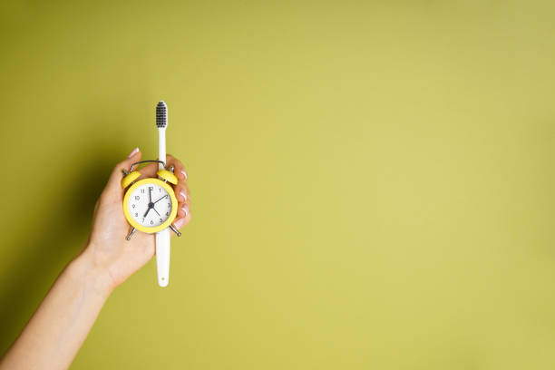 Yellow alarm clock and a toothbrush in the hand of a young woman on a green background, space fo text. Top view. Morning concept stock photo