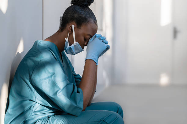 피곤한 우울한 여성 아프리카 스크럽 간호사가 병원 바닥에 얼굴 마스크 블루 유니폼 장갑을 끼고 있습니다. 소진 된 슬픈 검은 의사는 직장에서 코로나 바이러스 최전선 보호기도의 소진 스트� - consoling depression assistance 20s 뉴스 사진 이미지